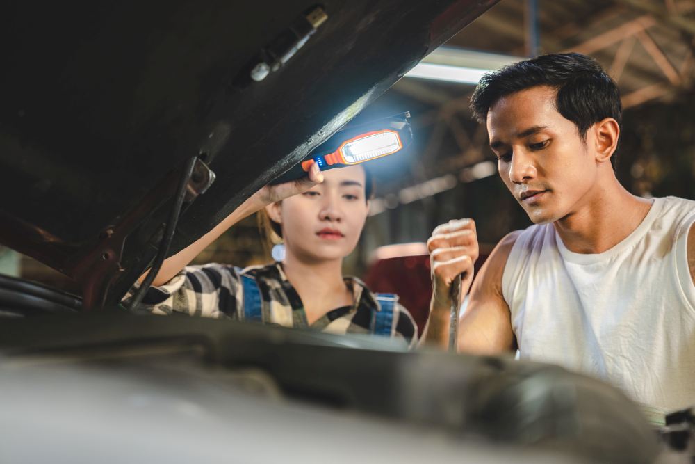 Two mechanics, one male and one female, work under the hood of a car using a handheld work light in a garage that specializes in used van sales Singapore.