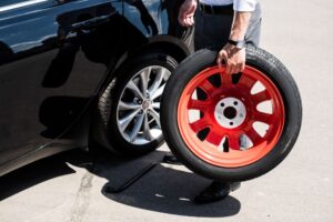 A person holds a red GTX02 spare tire next to a black car with one tire removed, preparing to change it on a sunny day.