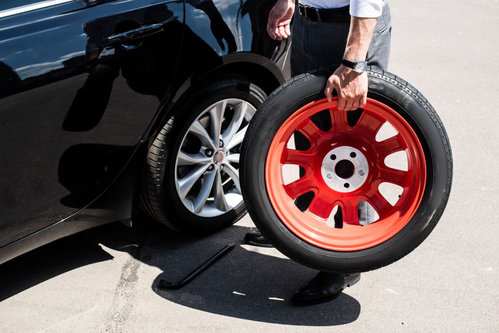 A person holds a red GTX02 spare tire next to a black car with one tire removed, preparing to change it on a sunny day.