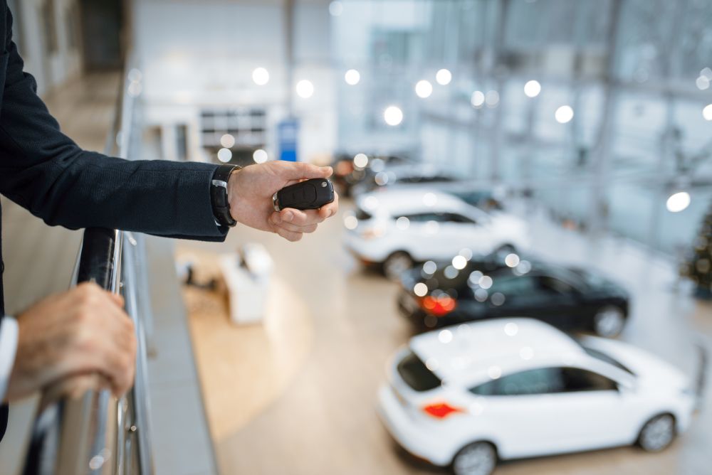 A person holding a car key fob overlooks several new cars displayed inside a modern, well-lit car showroom.