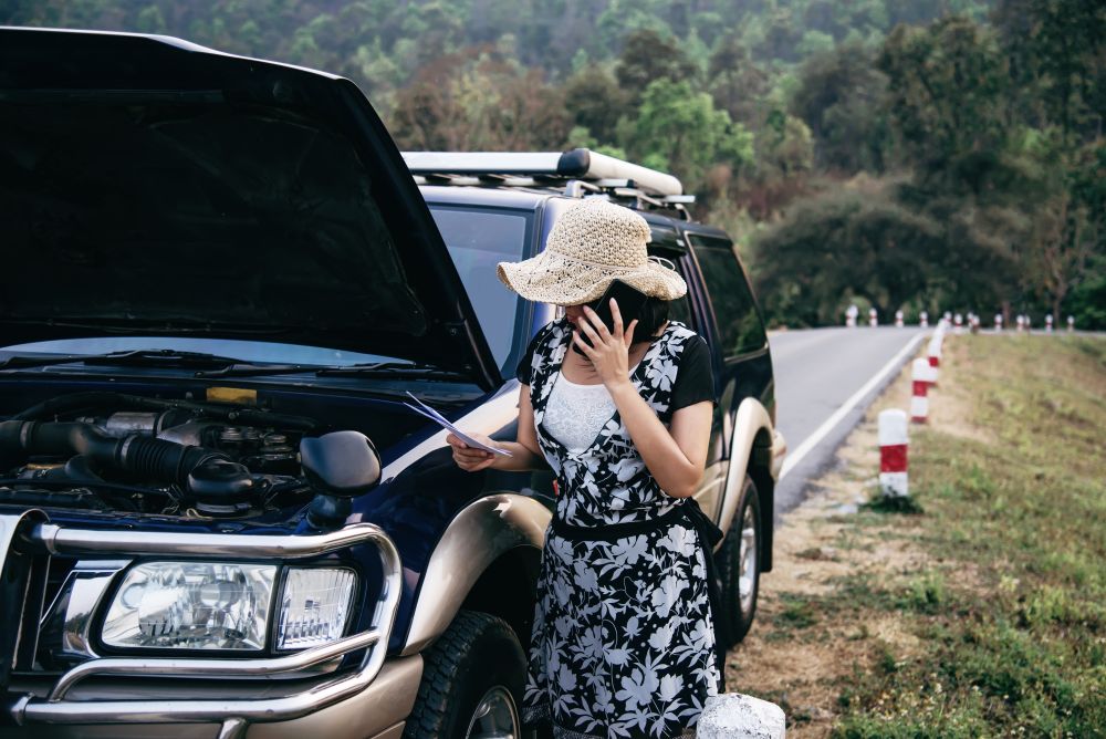 A person stands by a car with the hood open, looking at papers and talking on the phone beside a rural road, perhaps seeking advice on Scrap Car Singapore: What Vehicle Owners Should Know.