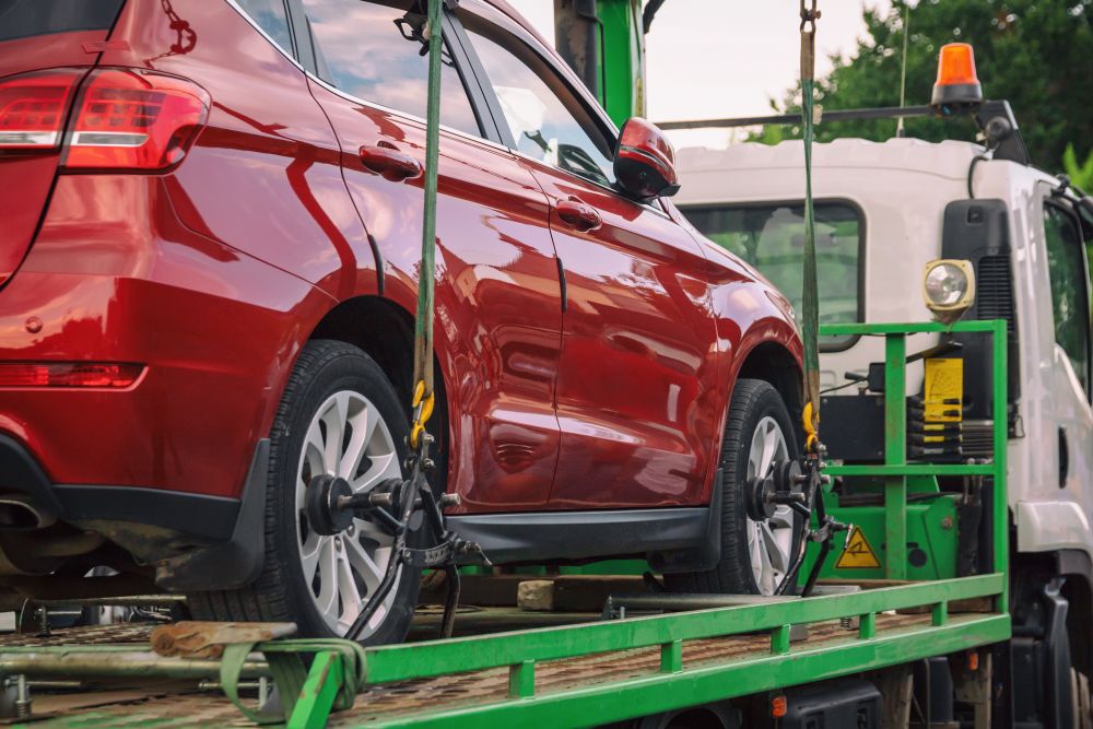 A red car is secured on a flatbed tow truck with metal straps, ready for transport—a clear example of when to use car towing services. Another vehicle is partially visible in the background.